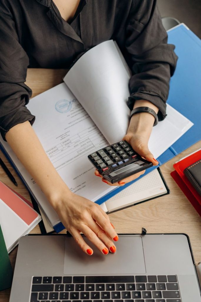 Maria Semple Bookkeeping A business professional uses a calculator among documents and laptop on a desk for financial tasks.